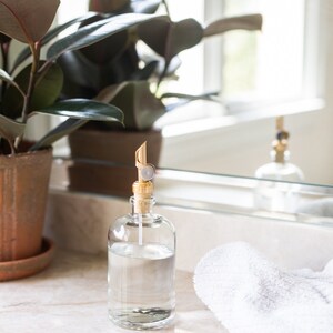May include: A clear glass bottle with a gold pump dispenser filled with liquid soap. The bottle is sitting on a white countertop next to a white towel.