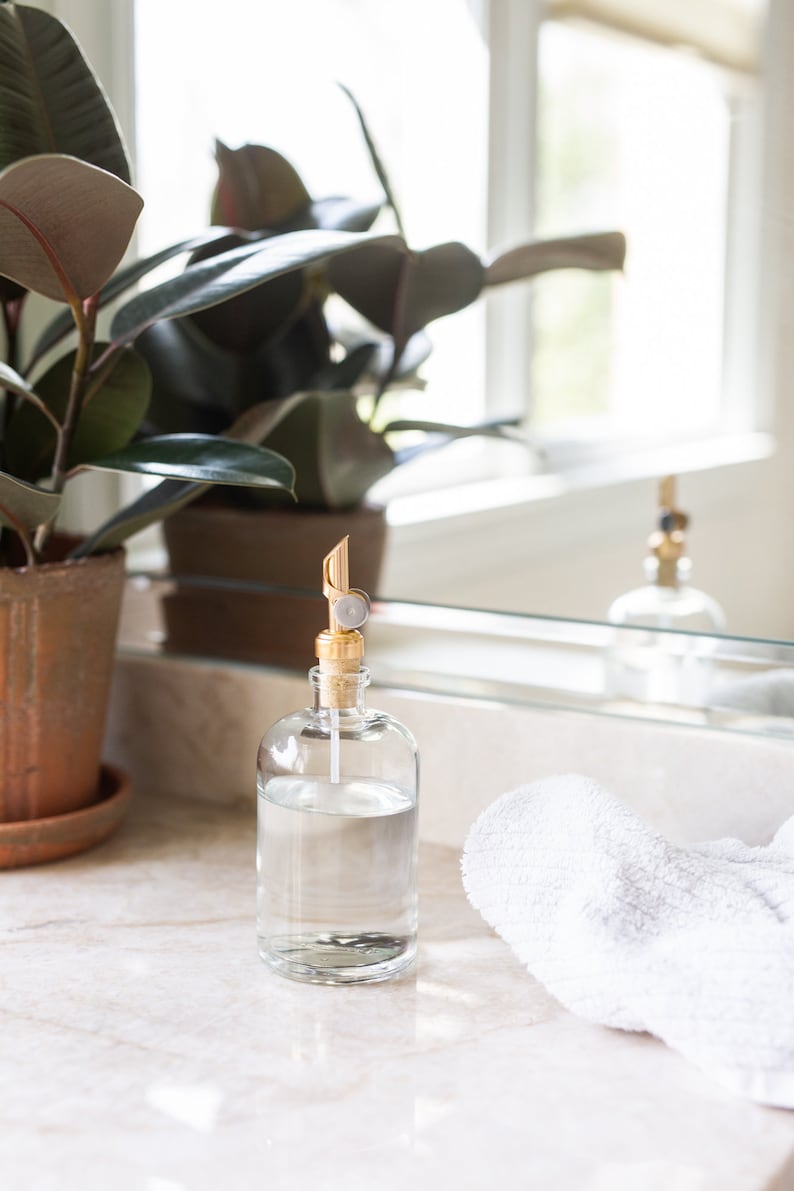 May include: A clear glass bottle with a gold pump dispenser filled with liquid soap. The bottle is sitting on a white countertop next to a white towel.