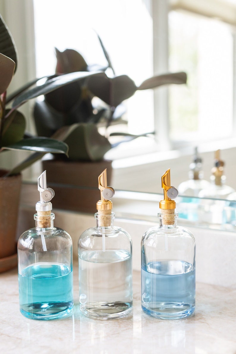 May include: Three glass bottles with gold and silver pump tops filled with blue liquid. The bottles are sitting on a white countertop.