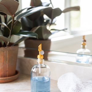 May include: A clear glass bottle with a blue liquid inside and a gold pump top. The bottle is sitting on a white countertop next to a white towel.