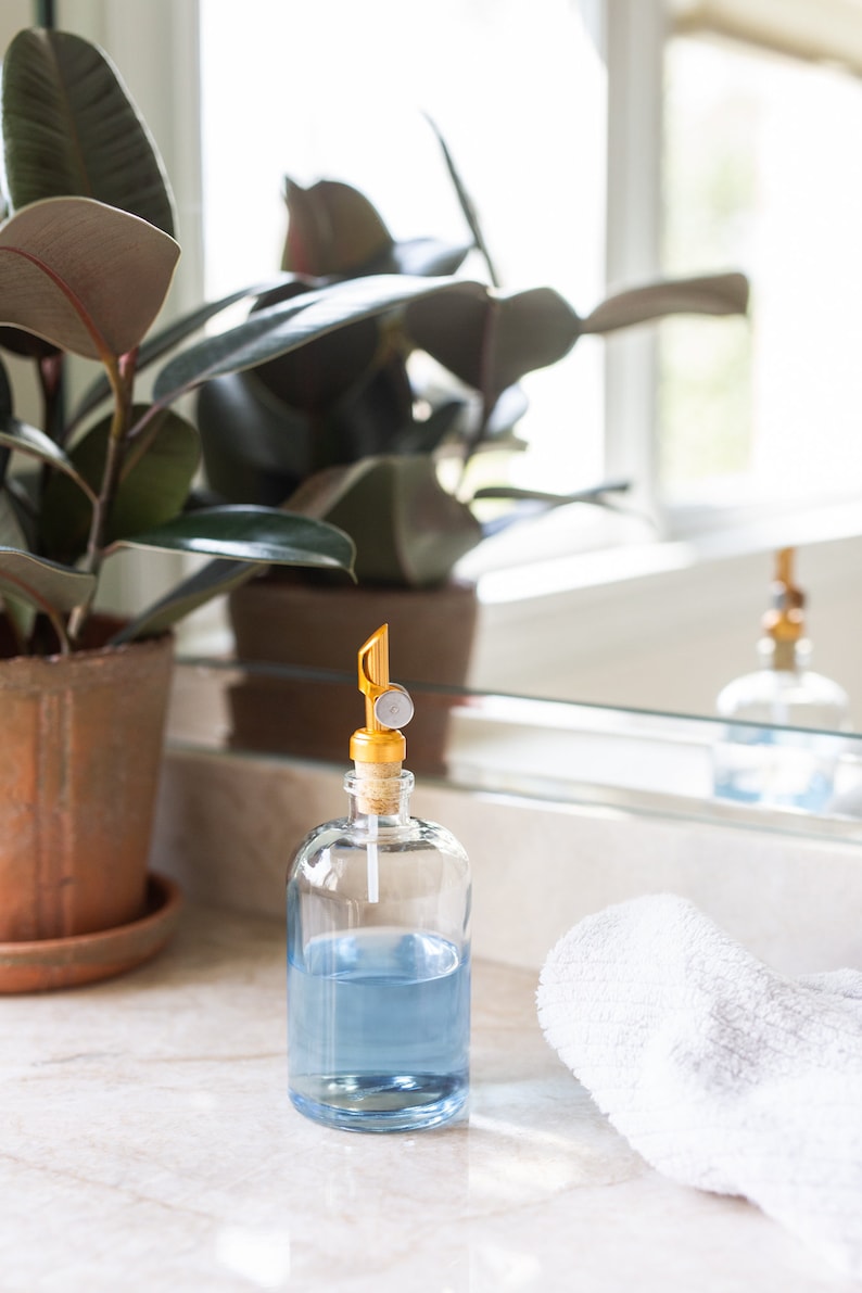 May include: A clear glass bottle with a blue liquid inside and a gold pump top. The bottle is sitting on a white countertop next to a white towel.