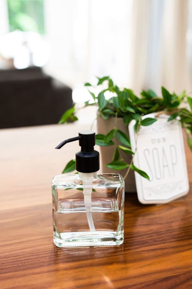 May include: A clear glass soap dispenser with a black pump top. The dispenser is filled with clear liquid and is sitting on a wooden surface. The word "SOAP" is visible on a white sign in the background.