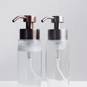 May include: Two glass soap dispensers with pumps. The dispenser on the left has a copper-colored pump and the dispenser on the right has a silver-colored pump.