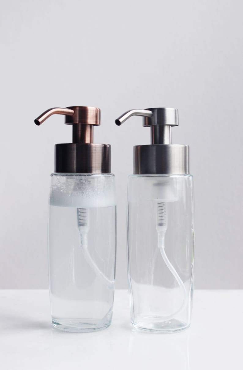 May include: Two glass soap dispensers with pumps. The dispenser on the left has a copper-colored pump and the dispenser on the right has a silver-colored pump.