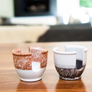 May include: Two ceramic cups with a unique design. The cup on the left is white with a brown speckled pattern. The cup on the right is white with a black speckled pattern. Both cups have a cut-out design on the side.