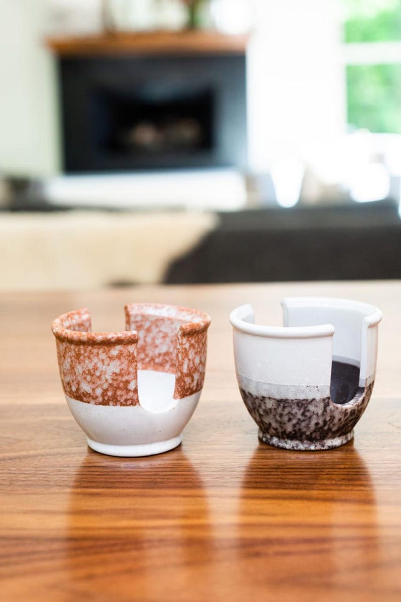 May include: Two ceramic cups with a unique design. The cup on the left is white with a brown speckled pattern. The cup on the right is white with a black speckled pattern. Both cups have a cut-out design on the side.
