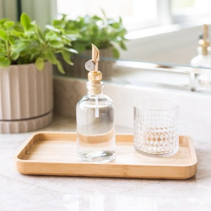 May include: A clear glass bottle with a gold pump top and a cork stopper. The bottle is filled with a clear liquid and is sitting on a wooden tray with a clear glass tumbler next to it.