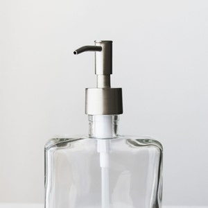 May include: A clear glass soap dispenser with a silver pump. The dispenser has a square shape and a wide mouth.