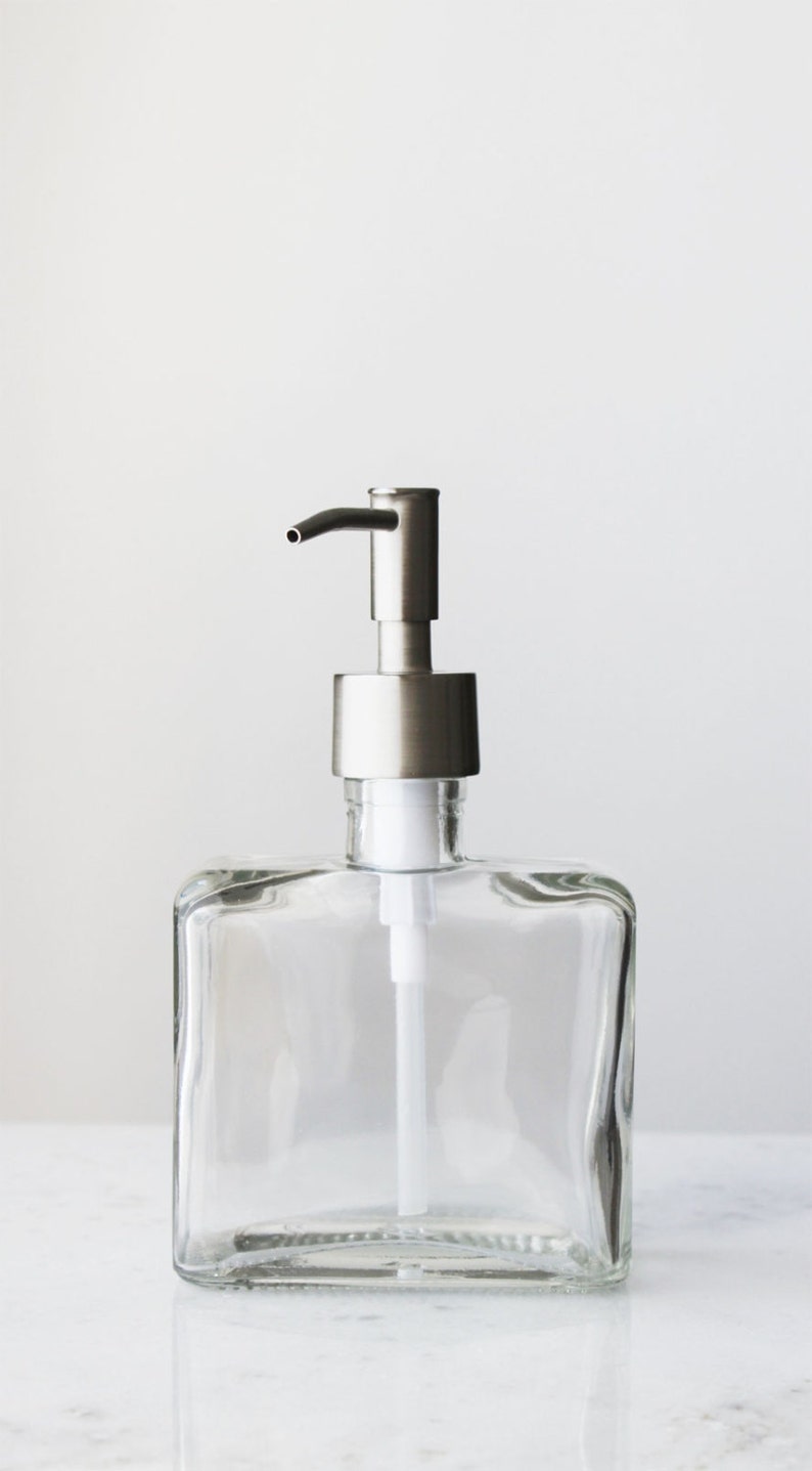 May include: A clear glass soap dispenser with a silver pump. The dispenser has a square shape and a wide mouth.