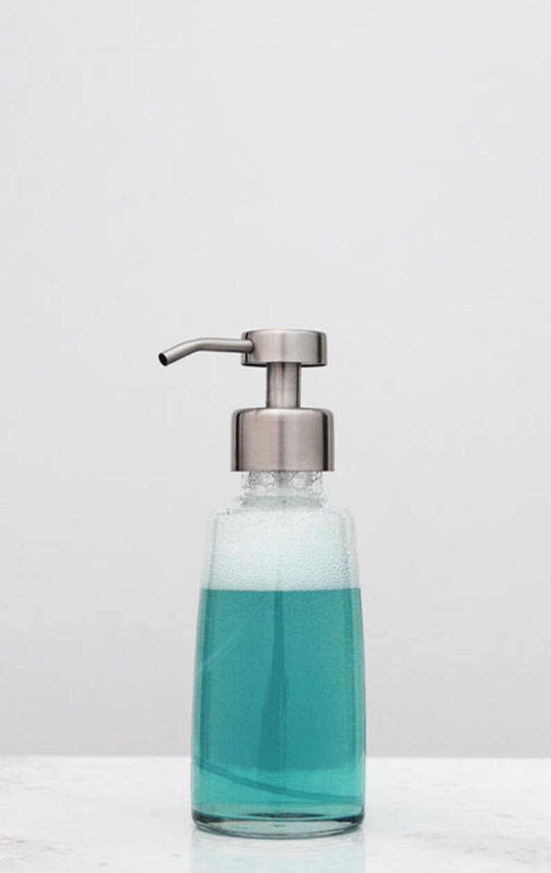 May include: A stainless steel soap dispenser with a clear glass bottle filled with blue liquid and white foam.