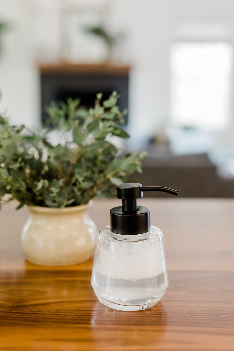 K&ouml;nnte beinhalten: Ein durchsichtiger Seifenspender aus Glas mit einer schwarzen Pumpe und einer wei&szlig;en, schaumigen Fl&uuml;ssigkeit im Inneren. Der Spender steht auf einer Holzoberfl&auml;che.