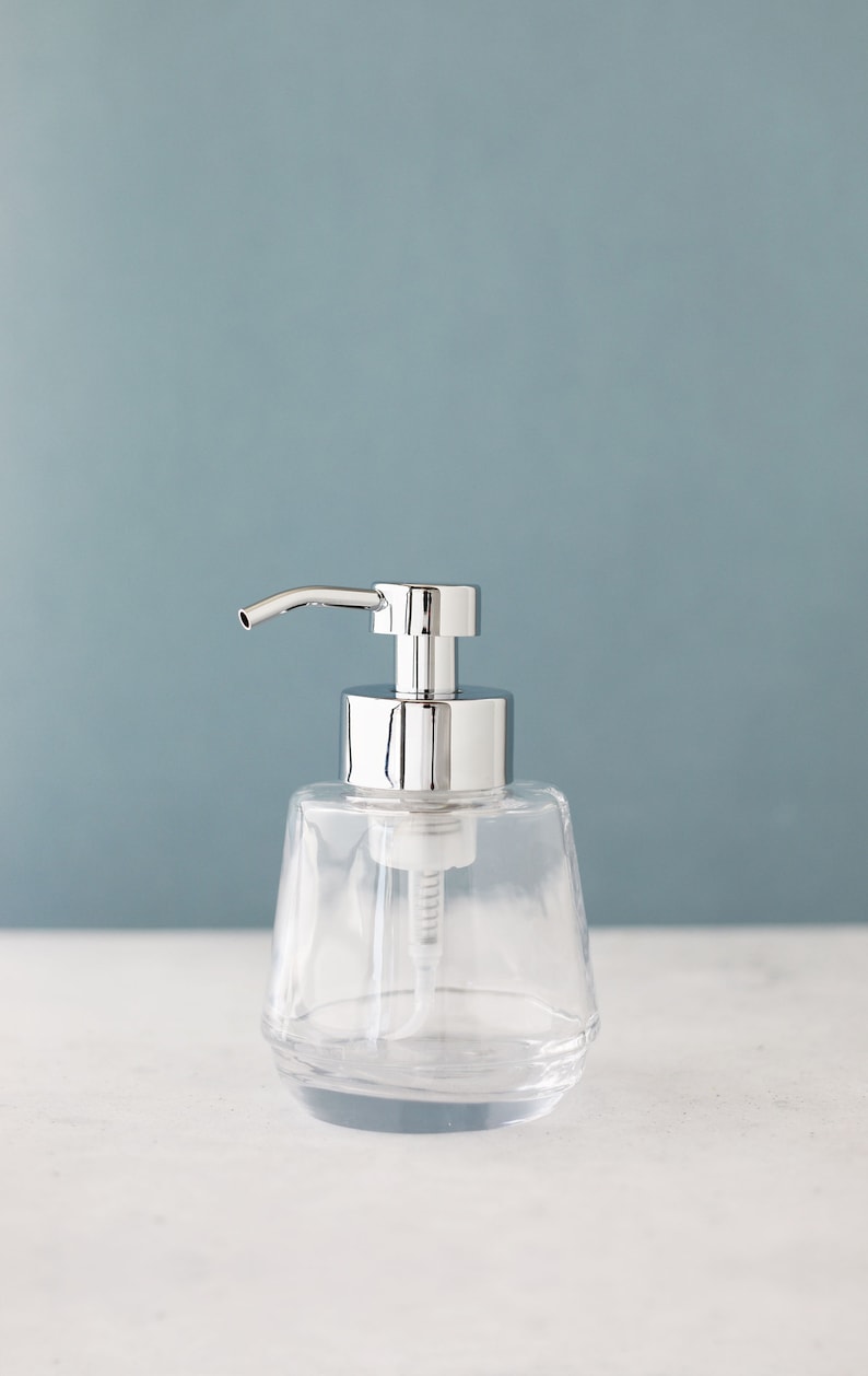 Pu&ograve; includere: Un dispenser di sapone in vetro trasparente con una pompa argentata. Il dispenser ha una base arrotondata e testurizzata.