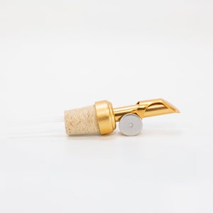 May include: Gold bottle pourer with a cork stopper and a metal spout. The spout is designed to control the flow of liquid.