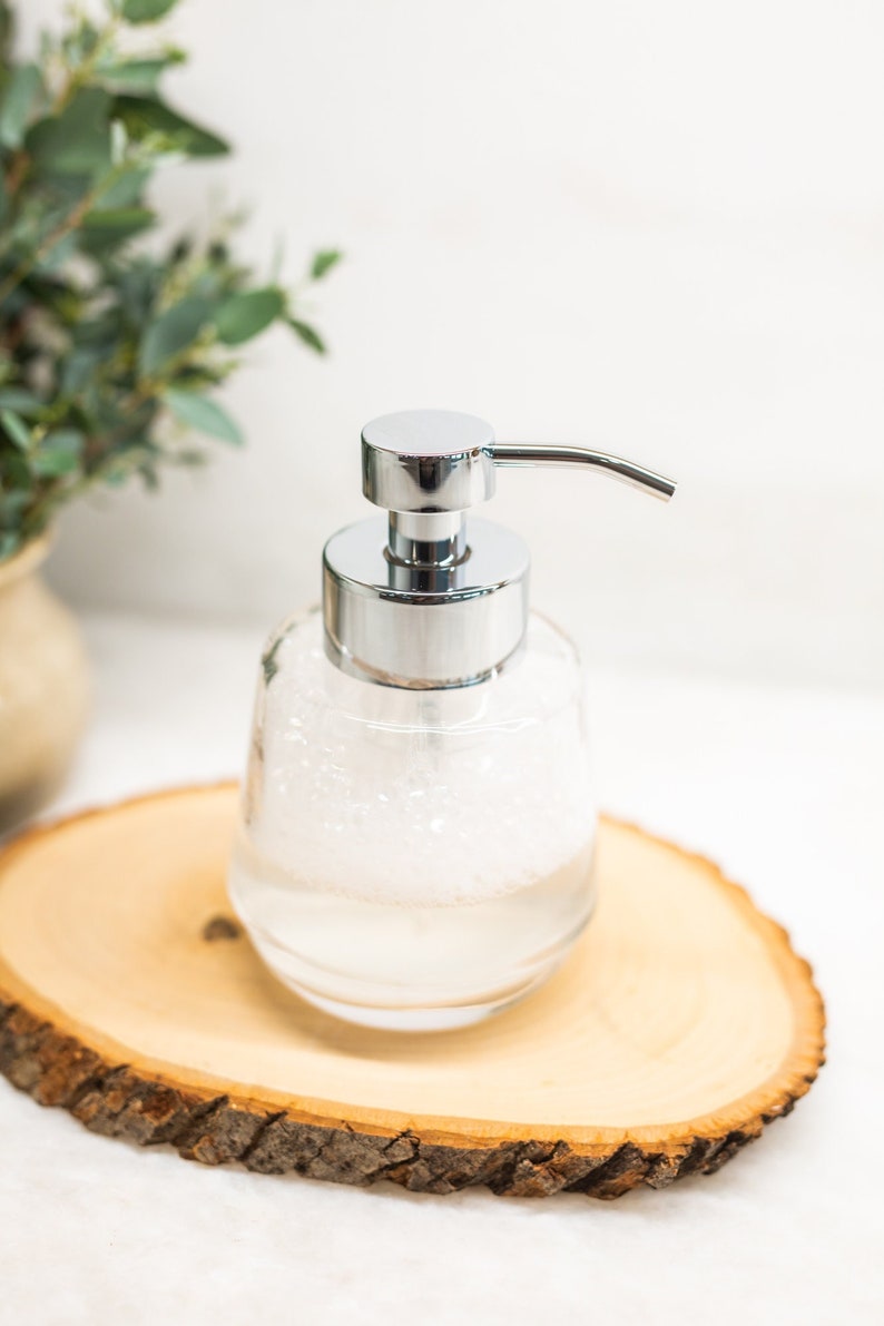 Pu&ograve; includere: Un dispenser di sapone in vetro trasparente con una pompa cromata e un coperchio argentato. Il dispenser &egrave; riempito con un liquido schiumoso bianco e si trova su una fetta di legno.