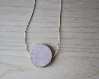 May include: A simple wooden pendant necklace with a round, light brown wooden disc on a thin, light beige cord.