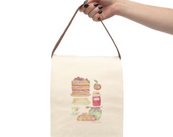 May include: Cream-colored tote bag with a brown handle, featuring a watercolor illustration of a layered cake, apple, jam, butter, tea cup, and bread. The bag is held by a person.