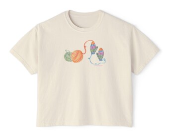 May include: Cream-colored t-shirt featuring a colorful illustration of yarn balls and mittens. The design includes green, orange, and blue yarn balls, with the mittens in shades of blue, green, and orange. The shirt has a crew neck and short sleeves.