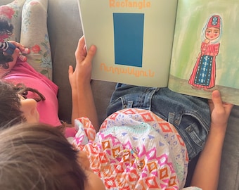 May include: Two children are reading a book together. The book is open to a page with a blue rectangle and the word "Rectangle" written in orange. The other page shows a drawing of a woman wearing a colorful dress and a headscarf. The text on the page is in Armenian.