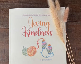 May include: A guided journal titled "Loving Kindness" with a watercolor illustration of yarn and mittens. The cover is cream-colored with text in orange and red. Dried ornamental grass is in the background.