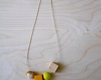 May include: A handmade necklace featuring a white cord with wooden beads. The necklace includes a gold and white marbled sphere, a yellow square, a green sphere, and a natural wood square. The beads are strung in a linear arrangement.