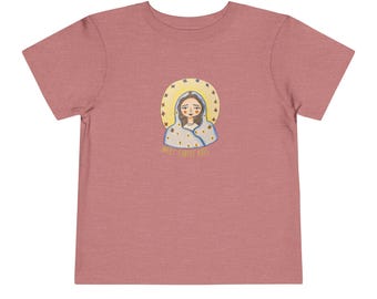 May include: A short-sleeved, dusty rose-colored t-shirt featuring a graphic of a woman with a halo and the text "MEET CHRIST KIDS". The illustration is in shades of blue, yellow, and white.