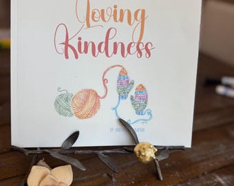 May include: A guided journal titled "Loving Kindness" with a watercolor illustration of yarn and mittens. The journal is displayed on a decorative metal stand with a flower accent. The cover text reads "A Guided Journal for the Highly Sensitive Christian Mama."