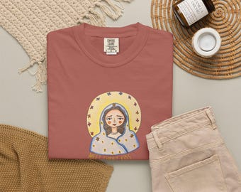 May include: A dusty rose-colored t-shirt featuring a hand-drawn illustration of a woman with a halo. The illustration is surrounded by a yellow halo with blue stars. The text "MEET CHRIST KIDS" is below the illustration. The shirt is paired with tan pants.