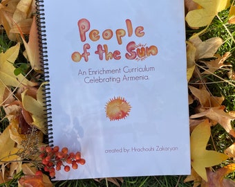 May include: A white spiral bound book with the title "People of the Sun" in colorful, stylized lettering. The subtitle reads "An Enrichment Curriculum Celebrating Armenia." The book is lying on a bed of green grass and autumn leaves.