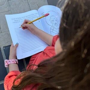 May include: A young person is coloring a black and white illustration of a saint in a coloring book. The book has a lined page with the text "Live simply, love generously, care deeply for the needs of your neighbor, speak softly... And leave the God." The person is using a yellow pencil with a pink eraser.