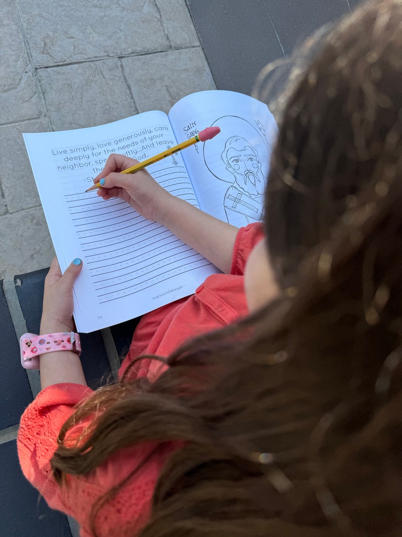 May include: A young person is coloring a black and white illustration of a saint in a coloring book. The book has a lined page with the text "Live simply, love generously, care deeply for the needs of your neighbor, speak softly... And leave the God." The person is using a yellow pencil with a pink eraser.