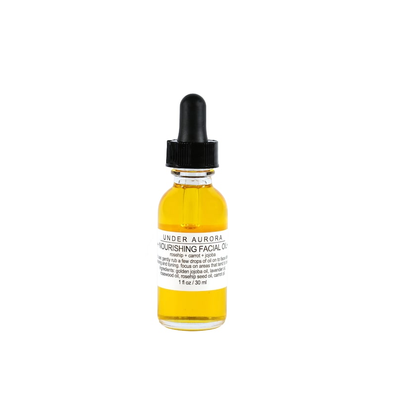 K&ouml;nnte beinhalten: Eine klare Glasflasche Under Aurora Nourishing Facial Oil mit einer schwarzen Pipette. Die Flasche enth&auml;lt eine goldgelbe Fl&uuml;ssigkeit. Das Etikett enth&auml;lt den Text "rosehip + carrot + jojoba" und "1 fl oz / 30 ml".