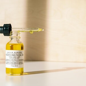 K&ouml;nnte beinhalten: Eine kleine Glasflasche Under Aurora Nourishing Facial Oil mit einer schwarzen Pipette. Die Flasche ist mit einer gelben Fl&uuml;ssigkeit gef&uuml;llt, und ein Tropfen &Ouml;l h&auml;ngt an der Pipette. Das Etikett enth&auml;lt den Text "1 fl oz | 30 ml".