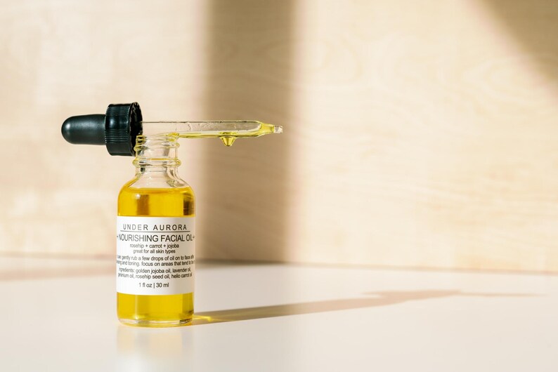 K&ouml;nnte beinhalten: Eine kleine Glasflasche Under Aurora Nourishing Facial Oil mit einer schwarzen Pipette. Die Flasche ist mit einer gelben Fl&uuml;ssigkeit gef&uuml;llt, und ein Tropfen &Ouml;l h&auml;ngt an der Pipette. Das Etikett enth&auml;lt den Text "1 fl oz | 30 ml".