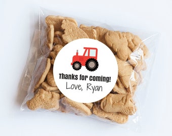 May include: A clear plastic bag filled with animal-shaped crackers. A white circular sticker with a red tractor graphic and the text "Thanks for coming! Love, Ryan" is affixed to the bag. The crackers are light brown.