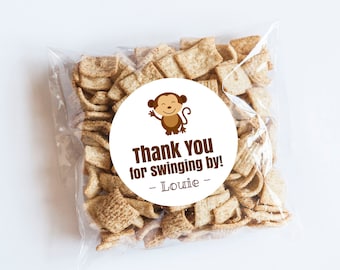 Puede incluir: Una pegatina circular blanca con una ilustración de un mono marrón y el texto "¡Gracias por pasar! - Lourie -" impreso en ella. La pegatina está en una bolsa de plástico transparente llena de pequeños trozos de cereal crujiente marrón.