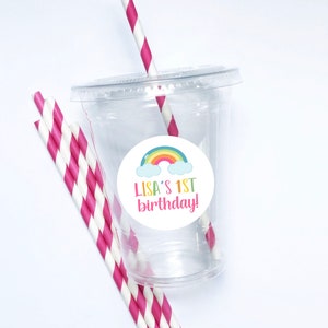 May include: A clear plastic cup with a pink and white striped straw. A white circle sticker with a rainbow and clouds is attached to the cup. The sticker says "LISA'S 1ST birthday!"
