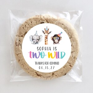 May include: A large, round cookie in a clear plastic wrapper. The cookie has a colorful circular label with the text "SOPHIA IS TWO WILD" and illustrations of an elephant, giraffe, and monkey. The text "THANKS FOR COMING! 06.15.27" is also included.