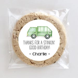 Op de afbeelding: Een ronde chocolate chip cookie in een doorzichtige cellofaan zak. De cookie heeft een witte ronde sticker met een groene illustratie van een vuilniswagen. De sticker leest: "THANKS FOR A STINKIN' GOOD BIRTHDAY! - Charlie".