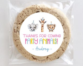 May include: A large, round chocolate chip cookie in a clear cellophane bag. The cookie has a white circular sticker with watercolor illustrations of an elephant, giraffe, and lion. Text reads "THANKS FOR COMING PARTY ANIMALS!" and "- Audrey -."