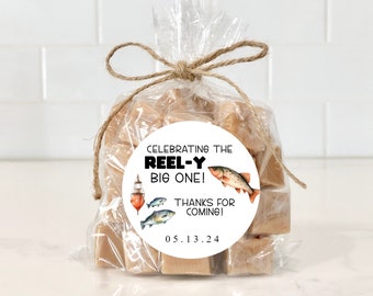 May include: A white circle sticker with black text that reads "Celebrating the Reel-Y Big One! Thanks for coming! 0.5.1.3.2.4" and a cartoon image of a fishing bobber and two fish. The sticker is attached to a clear plastic bag filled with small, square pieces of fudge.