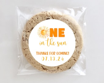 May include: A chocolate chip cookie in a clear cellophane bag. The cookie has a white sticker with orange text that reads "ONE in the sun" and "THANKS FOR COMING! 07.13.24". The sticker features a watercolor sun illustration.