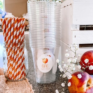 Puede incluir: Una pila de vasos de pl&aacute;stico transparente con una pegatina con tem&aacute;tica de melocot&oacute;n que dice "one sweet peach" en la parte delantera. Tambi&eacute;n hay pajitas rayadas de color naranja y blanco en un tarro de vidrio y melocotones en una caja de madera blanca.