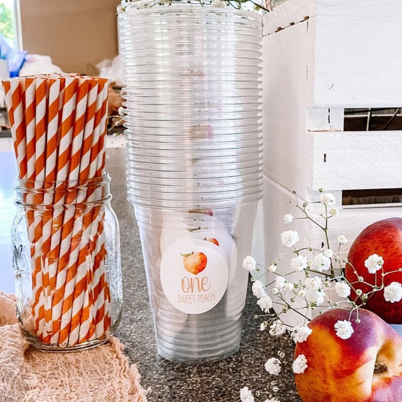 Puede incluir: Una pila de vasos de pl&aacute;stico transparente con una pegatina con tem&aacute;tica de melocot&oacute;n que dice "one sweet peach" en la parte delantera. Tambi&eacute;n hay pajitas rayadas de color naranja y blanco en un tarro de vidrio y melocotones en una caja de madera blanca.