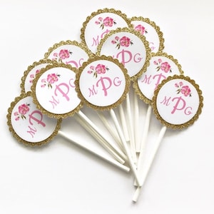 May include: A set of 10 cupcake toppers with a gold glitter background and pink floral design. Each topper features the initials "M P G" in pink lettering.