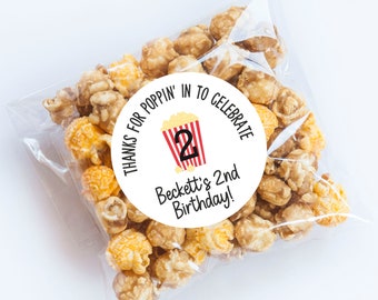 May include: Round white sticker with black text that says "Thanks for poppin' in to celebrate Beckett's 2nd birthday!"  A red and white striped popcorn bucket with the number 2 on it is in the center of the sticker.