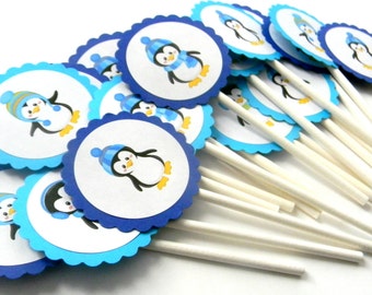 Pode incluir: Um conjunto de toppers de cupcake de pinguim azul e branco com palitos brancos. Os pinguins usam chapéus e cachecóis azuis.