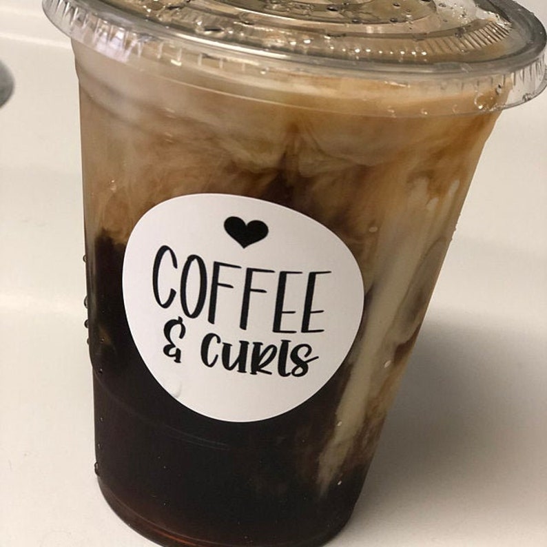 Puede incluir: Un vaso de pl&aacute;stico transparente lleno de una bebida de caf&eacute; helado marr&oacute;n y blanco. El vaso tiene una pegatina blanca con un coraz&oacute;n negro y el texto "COFFEE & curls".