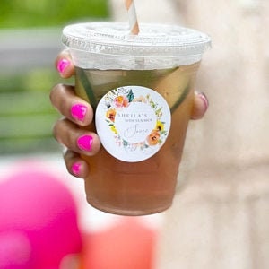 Puede incluir: Una pegatina redonda con un dise&ntilde;o de corona floral y el texto "SHEILA'S 30th SUMMER SOIREE" en un c&iacute;rculo blanco. La pegatina est&aacute; pegada a un vaso de pl&aacute;stico transparente con una pajita.