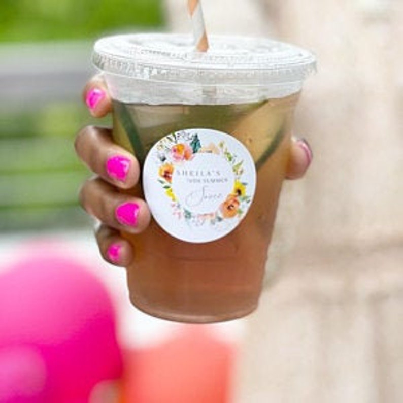 Puede incluir: Una pegatina redonda con un dise&ntilde;o de corona floral y el texto "SHEILA'S 30th SUMMER SOIREE" en un c&iacute;rculo blanco. La pegatina est&aacute; pegada a un vaso de pl&aacute;stico transparente con una pajita.