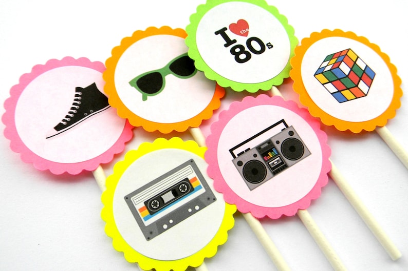 May include: Six colorful cupcake toppers with 80s themed designs. The toppers feature a cassette tape, a boombox, a pair of sunglasses, a Rubik's Cube, a high-top sneaker, and a heart with the text "I love the 80s".